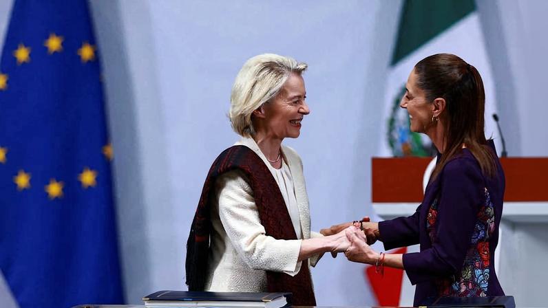 Duas mulheres sorriem e apertam as mãos em frente a uma mesa com documentos, com as bandeiras da União Europeia e do México ao fundo, em ambiente formal.