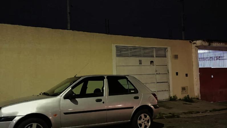 Carro prata estacionado na rua em frente a um muro amarelo com um portão branco e outro vermelho. Céu escuro ao fundo indica início da noite.