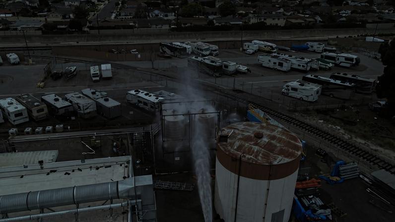 Vapor sai com força de um tanque cilíndrico branco com tampa enferrujada em área industrial. Ao redor, há veículos recreativos estacionados e trilhos de trem, com bairro residencial ao fundo.