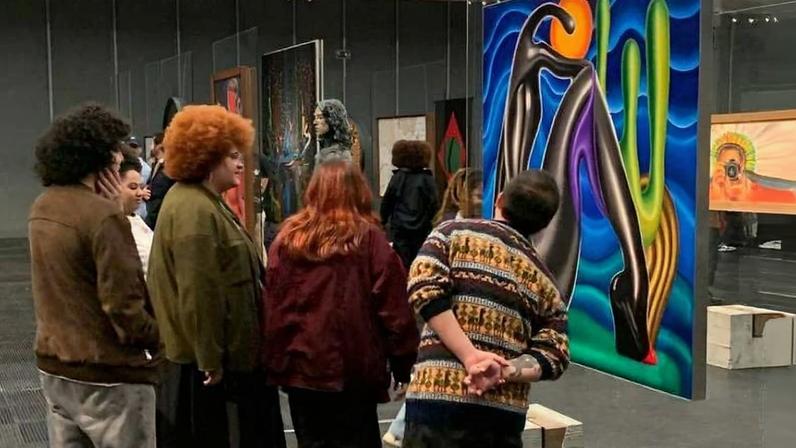 Seis pessoas em pé observam uma pintura abstrata colorida pendurada em uma galeria. A obra apresenta formas curvas em tons vibrantes de azul, verde, amarelo e roxo. O ambiente tem iluminação branca e paredes cinza, com bancos de madeira próximos.