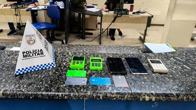No balcão de granito de uma delegacia, estão dispostos dois aparelhos de cartão verde, três cartões bancários, dois celulares e uma calculadora. Ao lado, há um suporte triangular com a inscrição 'Polícia Militar'. Ao fundo, duas pessoas trabalham em computadores em um ambiente de escritório.