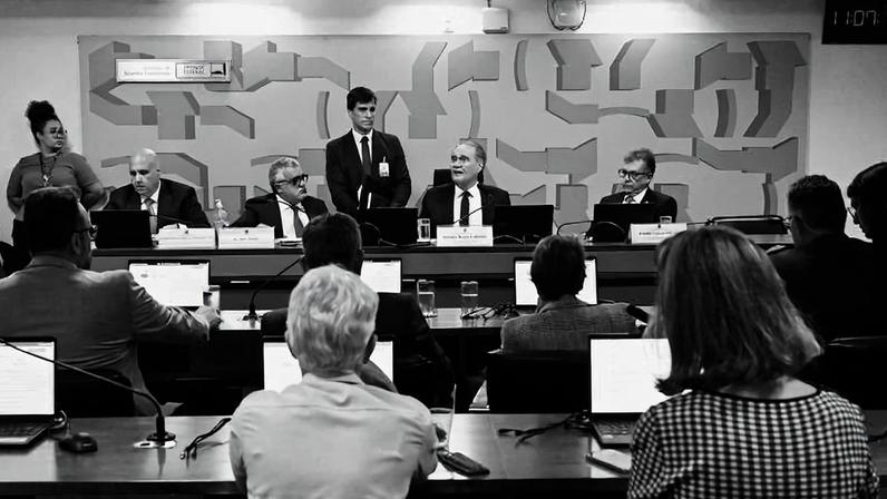Seis pessoas sentadas em mesa de reunião com laptops e microfones, cinco delas voltadas para a audiência. Um homem está em pé ao centro, falando. Ao fundo, parede com painel decorativo abstrato e relógio digital vermelho.