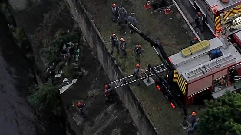 Bombeiros e equipes de resgate atendem acidente grave na Avenida do Estado próximo à Marginal Tietê. Caminhão de bombeiros está estacionado na via, com escada apoiada no muro ao lado do rio. Várias pessoas estão no local, incluindo socorristas e policiais, enquanto um carro branco está parado na pista ao lado.