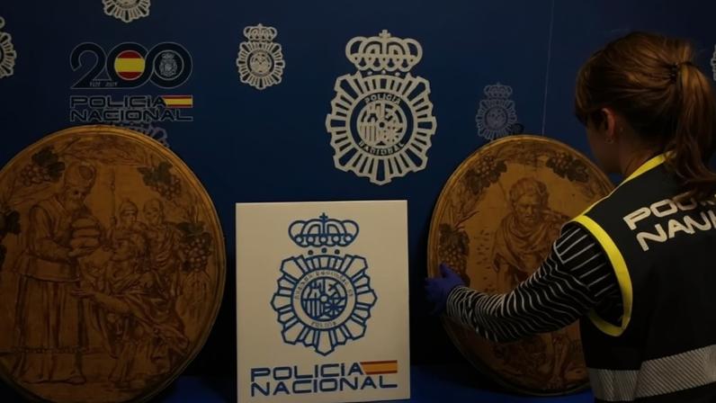Policial da Polícia Nacional, com colete preto e amarelo, examina dois objetos circulares grandes com gravuras antigas em tom marrom. Ao fundo, parede azul com emblemas da Polícia Nacional e logo comemorativo de 200 anos.