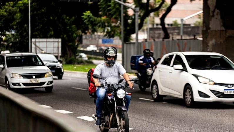 Motociclista usando capacete preto e camiseta cinza dirige motocicleta com mochila vermelha nas costas em avenida urbana. Ao redor, carros brancos e um motociclista ao fundo trafegam na mesma via, com árvores e construções ao fundo.