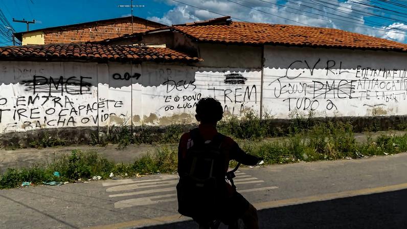 Pessoa com mochila sentada em bicicleta na sombra observa muro branco com várias pichações em preto. O muro tem telhado de cerâmica e está em área urbana com céu parcialmente nublado ao fundo.