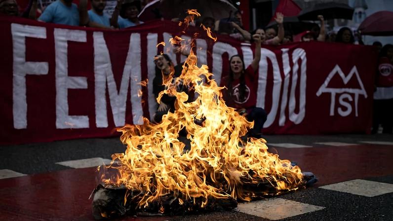 Grupo de manifestantes segura faixa vermelha com a palavra 'FEMINICÍDIO' em branco. Na frente, fogo intenso queima objetos no chão, em via pública.