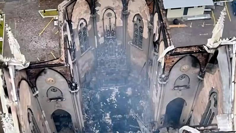 Vista aérea mostra interior de igreja com paredes queimadas e teto destruído. Resquícios de fumaça ainda sobem do local. Estruturas externas permanecem parcialmente intactas.