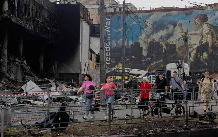 Sete pessoas caminham em calçada próxima a edifício parcialmente destruído com marcas de incêndio. Ao fundo, mural grande mostra cena de guerra com criança segurando estilingue e fumaça sobre prédios. Ambiente urbano com destroços e barreiras metálicas na frente.
