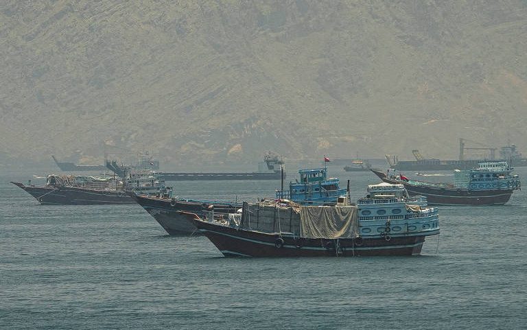 Quatro barcos tradicionais de madeira navegam em águas calmas próximas a uma costa montanhosa. As embarcações estão distribuídas em diferentes distâncias, com uma delas em primeiro plano e as outras três ao fundo, próximas à base das montanhas.