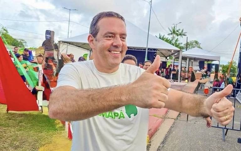 Homem de camiseta branca com logo verde sorri e faz gesto de positivo com as duas mãos em evento ao ar livre. Ao fundo, pessoas, tendas e equipamentos de som em área urbana com céu nublado.