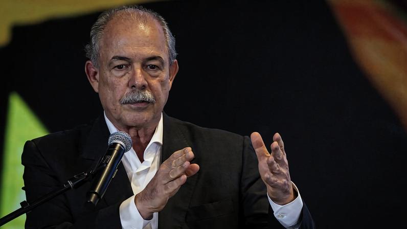 Homem de meia-idade com cabelo grisalho e bigode, vestindo terno preto e camisa branca, gesticula com as mãos enquanto fala ao microfone em um evento.