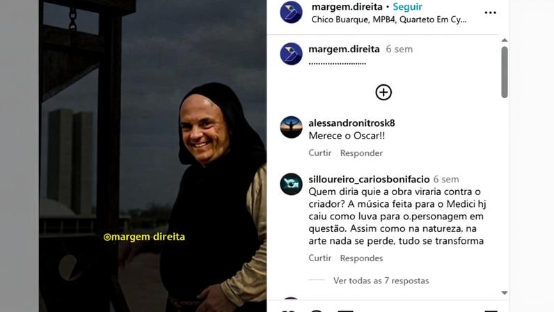 Homem sorridente vestido com hábito preto e túnica bege está ao lado de uma guilhotina de madeira em área externa. Ao fundo, edifícios altos e céu nublado.