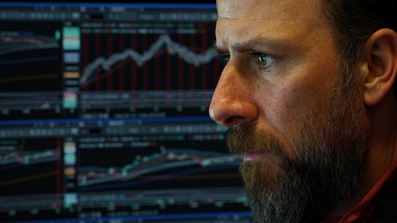 Homem de perfil com barba e expressão concentrada observa gráficos financeiros coloridos exibidos em várias telas ao fundo, mostrando linhas e barras de tendências de mercado.