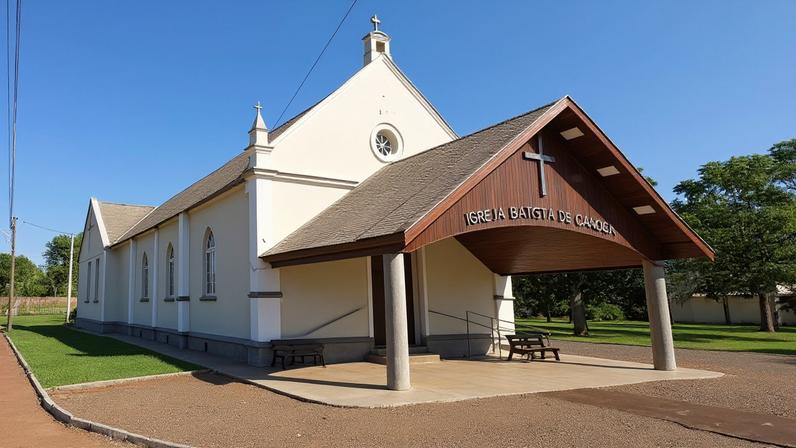 Igreja branca com telhado marrom e entrada coberta por alpendre de madeira. Vegetação ao redor inclui arbustos e árvores sob céu azul claro.