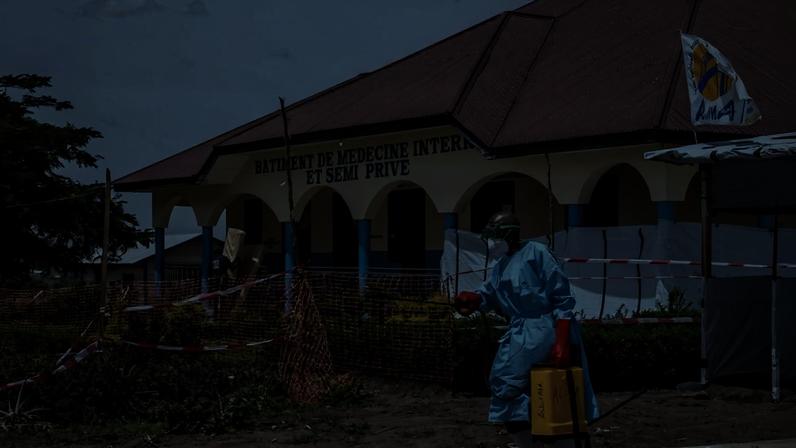 Pessoa vestindo equipamento de proteção completo, incluindo máscara, luvas vermelhas e avental azul, caminha em área externa próxima a prédio com placa 'Bâtiment de Médecine Interne et Semi Privé'. O local está cercado por barreiras de plástico laranja e fita de isolamento, com vegetação ao redor e céu parcialmente nublado.