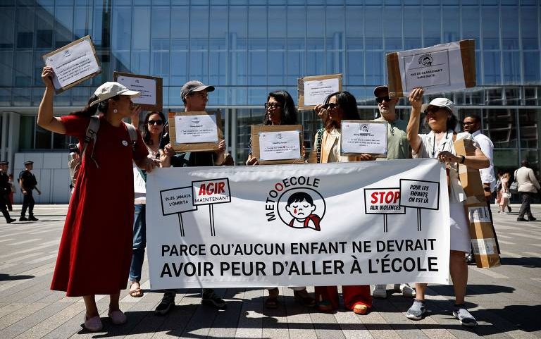 Oito pessoas em pé seguram uma faixa branca com texto em francês contra violência nas escolas, em frente a um prédio de vidro. Algumas seguram placas com mensagens relacionadas à causa.