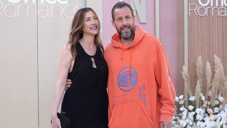 Homem e mulher posam juntos em frente a painel com o título 'Office Romance'. Ela veste vestido preto e ele usa moletom laranja com calça azul. Flores brancas decoram o ambiente.
