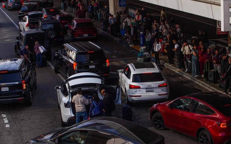 Diversos veículos parados em fila na via externa de terminal de aeroporto, com passageiros carregando bagagens e aguardando na calçada. Pessoas estão próximas aos carros, algumas abrindo porta-malas para guardar malas.