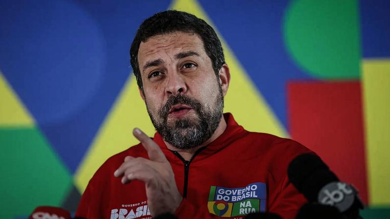 Homem de barba e cabelo curto veste jaqueta vermelha com logotipos do Governo do Brasil e fala em coletiva. Fundo colorido com formas geométricas em azul, amarelo, verde e vermelho. Microfones de várias emissoras estão posicionados à frente.