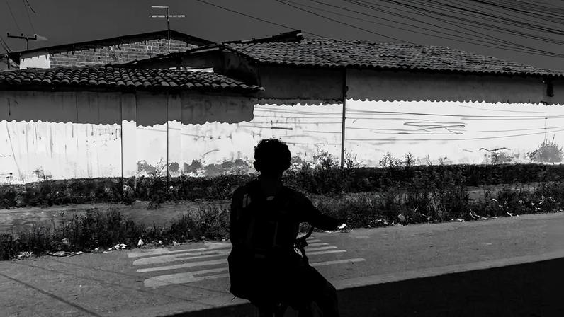 Pessoa com mochila sentada em bicicleta na sombra observa muro branco com várias pichações em preto. O muro tem telhado de cerâmica e está em área urbana com céu parcialmente nublado ao fundo.