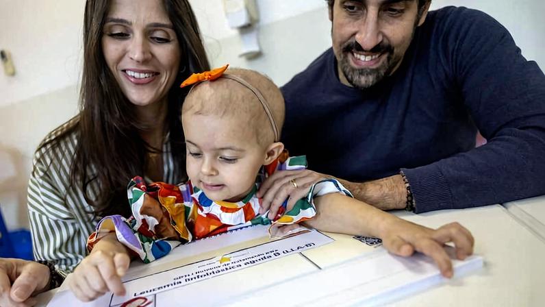 Criança pequena com faixa laranja na cabeça e vestido colorido toca material educativo sobre mesa branca, acompanhada por mulher e homem sorridentes ao fundo.