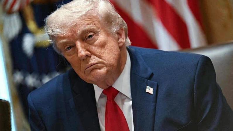 Presidente dos Estados Unidos sentado em cadeira, vestindo terno azul escuro, camisa branca e gravata vermelha. Bandeiras americanas desfocadas ao fundo.