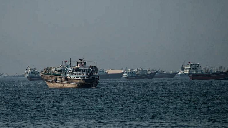 Dez navios de carga e pesca estão ancorados em mar calmo, distribuídos em linha irregular. O céu está nublado e sem sol visível, com horizonte pouco definido.