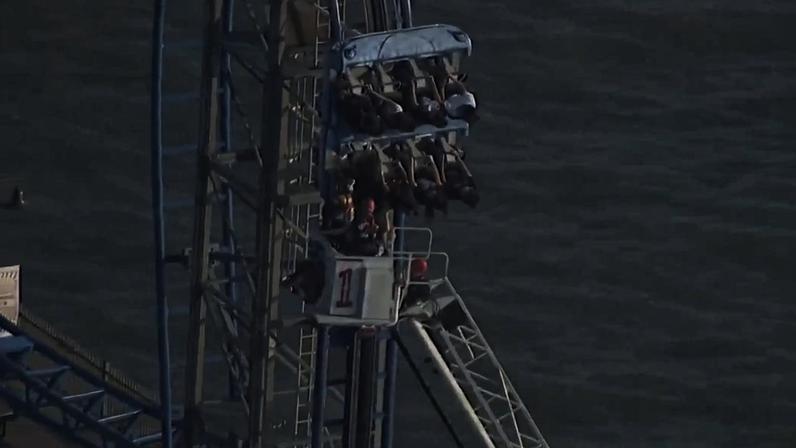 Passageiros em assentos suspensos de montanha-russa azul e branca, em curva elevada próxima a um corpo d'água. Estrutura metálica visível e céu claro.