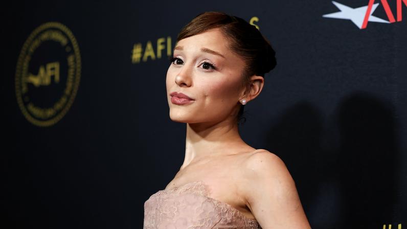 Mulher jovem com vestido tomara que caia bege posa de perfil em tapete vermelho com fundo preto e logos do AFI Awards.