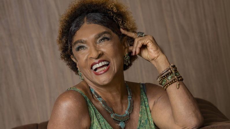 Mulher negra com vestido verde estampado sentada em poltrona de couro marrom, apoiando o braço direito no encosto e tocando a cabeça com a mão esquerda. Ela usa colares, pulseiras e anel, e está em ambiente interno com parede de madeira ao fundo.