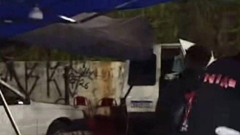 Homem de terno escuro está em área externa próxima a estação de trem, com destroços e objetos espalhados no chão. Veículo branco está estacionado ao fundo sob estrutura metálica.