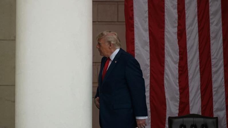 Homem de terno azul e gravata vermelha caminha ao lado de uma grande bandeira dos Estados Unidos pendurada verticalmente. Ele está de perfil, próximo a uma coluna branca, com uma cadeira de couro escuro ao fundo.