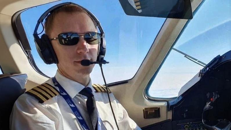 Homem vestido com uniforme de piloto, incluindo camisa branca com ombreiras douradas, gravata preta e fone de ouvido com microfone, sentado na cabine de comando de uma aeronave. Ele usa óculos escuros e está diante dos controles do avião, com céu azul visível pela janela.