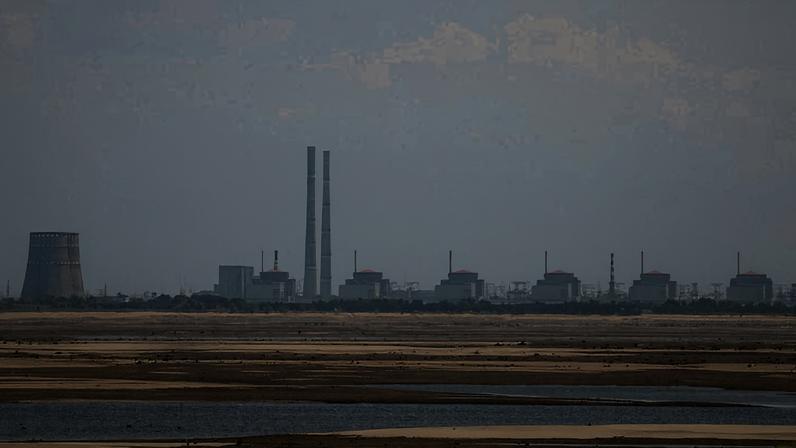 Complexo industrial com várias torres e chaminés ao fundo, visto além de uma extensa área alagada com bancos de areia e água. O céu está claro com poucas nuvens.
