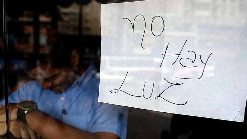 Papel branco com a frase 'No hay luz' escrita à mão está colado em vidro. Ao fundo, homem de camisa azul parece ajustar relógio ou pulseira, em ambiente interno pouco iluminado.