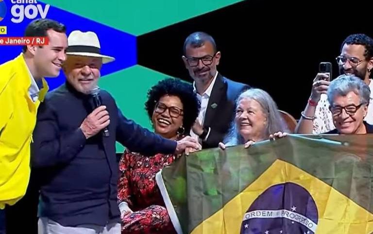 O presidente Lula (PT) segura bandeira do Brasil em evento no Rio de Janeiro neste sábado (30). Ele está ao lado do prefeito do Rio, Eduardo Cavaliere (PSD), com casaco da seleção brasileira e de artistas como o ator Paulo Betti.
