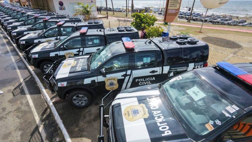 Diversas viaturas pretas e brancas da Polícia Civil estão estacionadas em fila dupla em área externa com vista para o mar. As viaturas possuem sirenes vermelhas e azuis no teto e estão alinhadas lado a lado em um espaço pavimentado próximo a palmeiras e faixa de areia. Ao fundo, há um grande balão branco com inscrições e um quiosque.