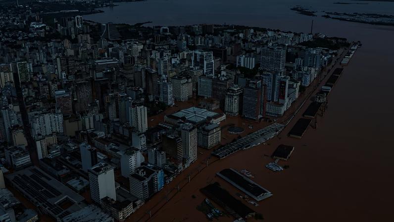 Vista aérea de Porto Alegre inundada, com prédios e ruas submersos em água barrenta. O centro da cidade e o cais do porto estão cobertos pela enchente, com apenas os telhados de algumas estruturas visíveis. O rio Guaíba, também marrom, se estende ao fundo, sob um céu nublado