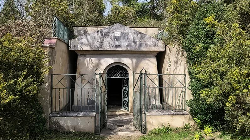 The entrance to a mausoleum. There is a green gate at the front, with plants surrounding the entrance.