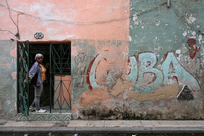 Moradora olha da porta de casa ao lado de um muro com a palavra "Cuba" pichada, em rua de Havana