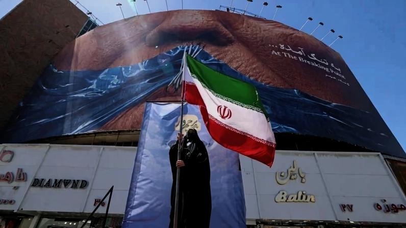 Uma mulher segura a bandeira iraniana enquanto está em frente a um outdoor anti-EUA que faz referência ao presidente Donald Trump e ao Estreito de Ormuz, instalado em um prédio na Praça Valiasr, em Teerã, em 10 de maio de 2026 - (crédito: ATTA KENARE / AFP)