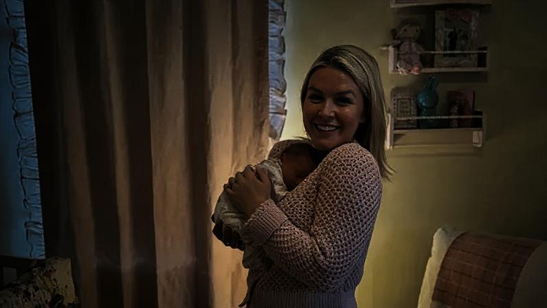 Karoline Leavitt via Instagram A young woman with blonde hair wears a pink knit sweater and smiles while holding a small baby on her chest. In the background is a white lounge chair with a pink blanket and the corner of a crib.