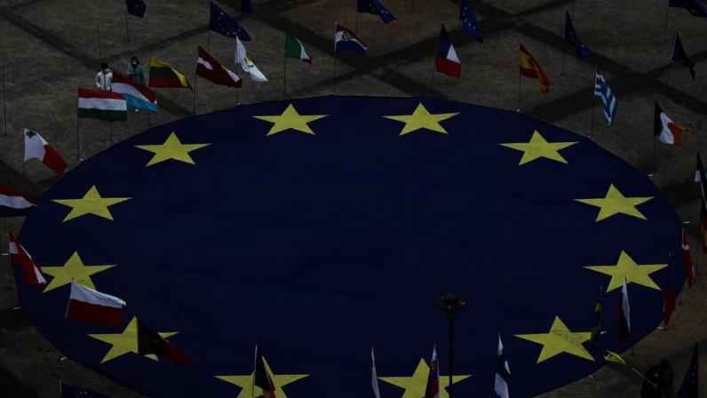 Enorme bandeira da União Europeia no chão, cercada por bandeiras dos países membros. Pessoas montando a exibição.