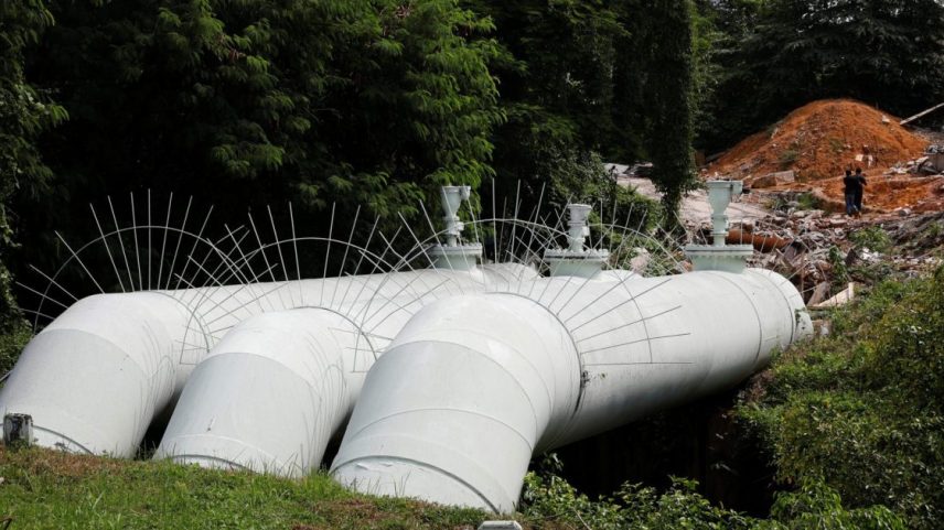 Grandes tubulações brancas deitadas na grama. Ao fundo, árvores e um monte de terra vermelha com duas pessoas.