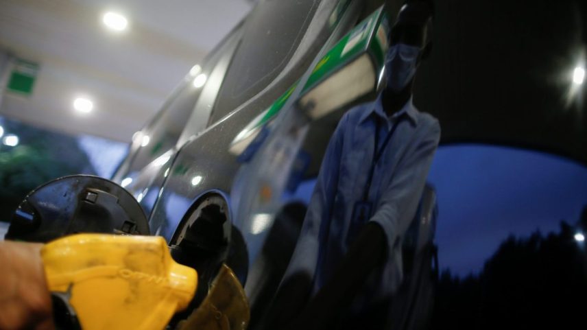 Pessoa abastece carro com bico amarelo. Frentista mascarado refletido na lataria escura do veículo.