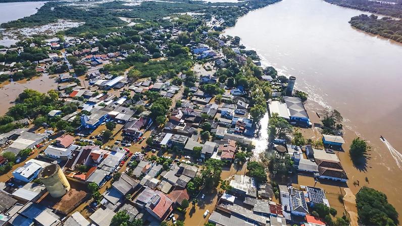 vista aérea de área residencial às margens de rio atingida por inundações, com as águas ocupando o leito das ruas por entre as casas, que se concentram na parte inferior da imagem, enquanto na superior aparecem áreas verdes tomadas pelas águas