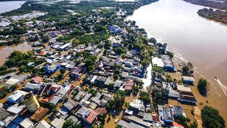 vista aérea de área residencial às margens de rio atingida por inundações, com as águas ocupando o leito das ruas por entre as casas, que se concentram na parte inferior da imagem, enquanto na superior aparecem áreas verdes tomadas pelas águas