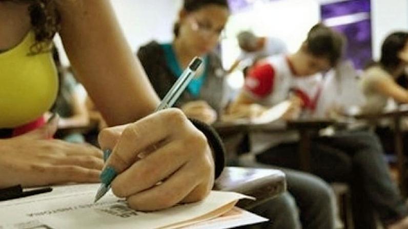 Pessas sentadas numa sala de aula fazendo uma prova e foco numa mão com uma caneta anotando num papel