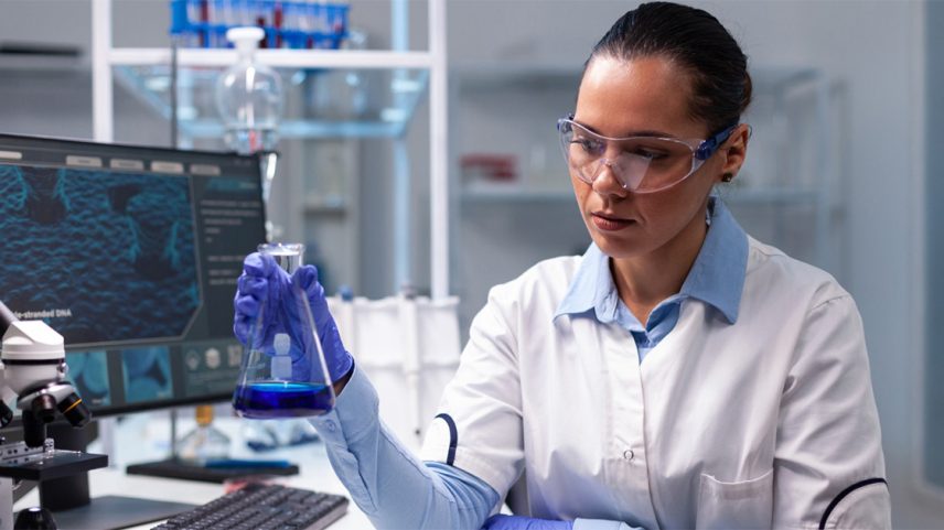Imagem de uma jovem pesquisadora, vestindo jaleco branco sobre camisa azul, manipulando um frasco em um laboratório, onde se encontram também um microscópico e um computador